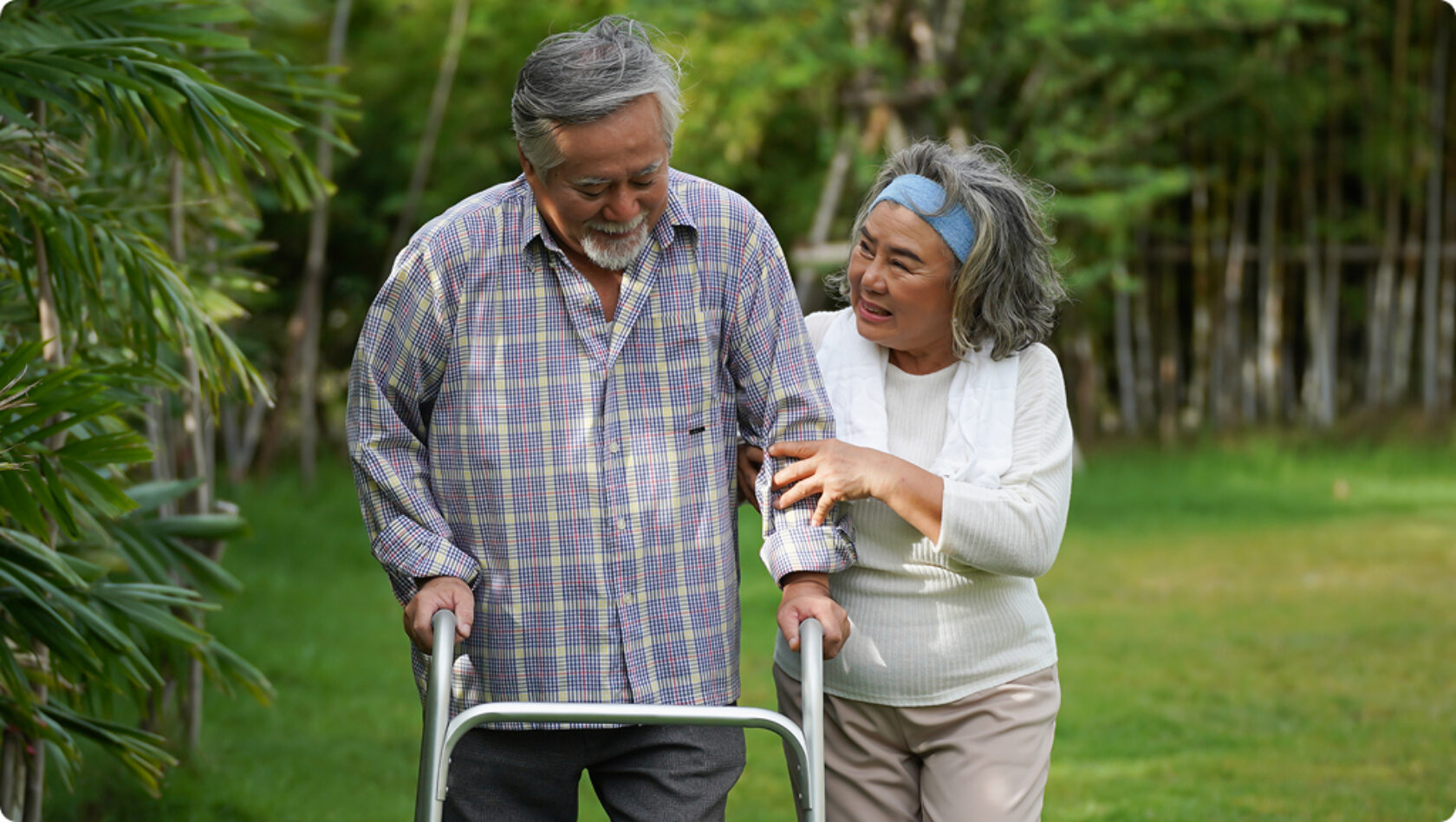 A woman supports a man using a walker during a walk outside.
